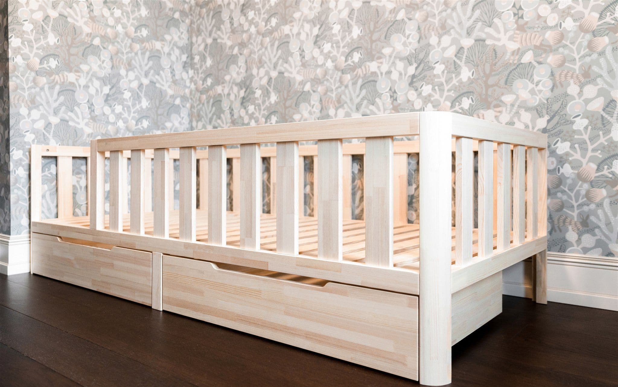 Natural solid wood Montessori toddler bed frame with safety guardrails, rounded corners and two pull-out storage drawers on a dark floor
